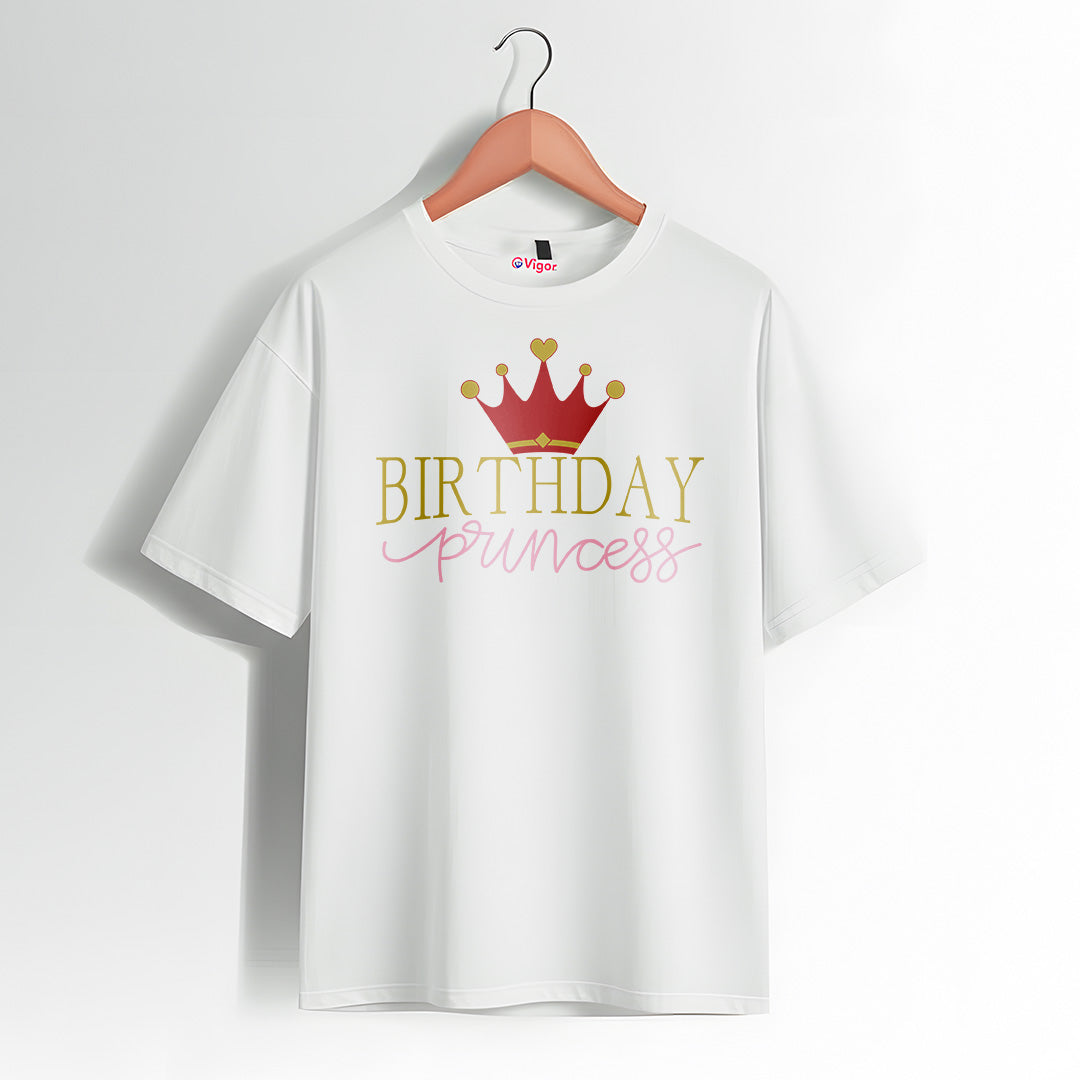 White T-shirt hanging on a wooden hanger with a design that reads “Birthday Princess” in gold and pink text, featuring a red and gold crown graphic above the text.