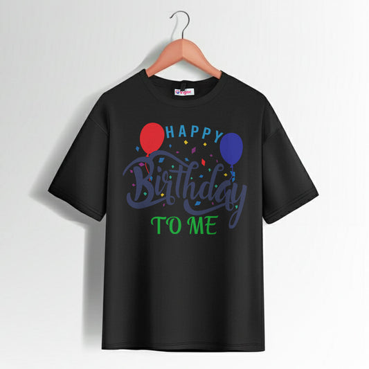 Black T-shirt with colorful “Happy Birthday To Me” text design, featuring red and blue balloons and confetti graphics, hanging on a wooden hanger.